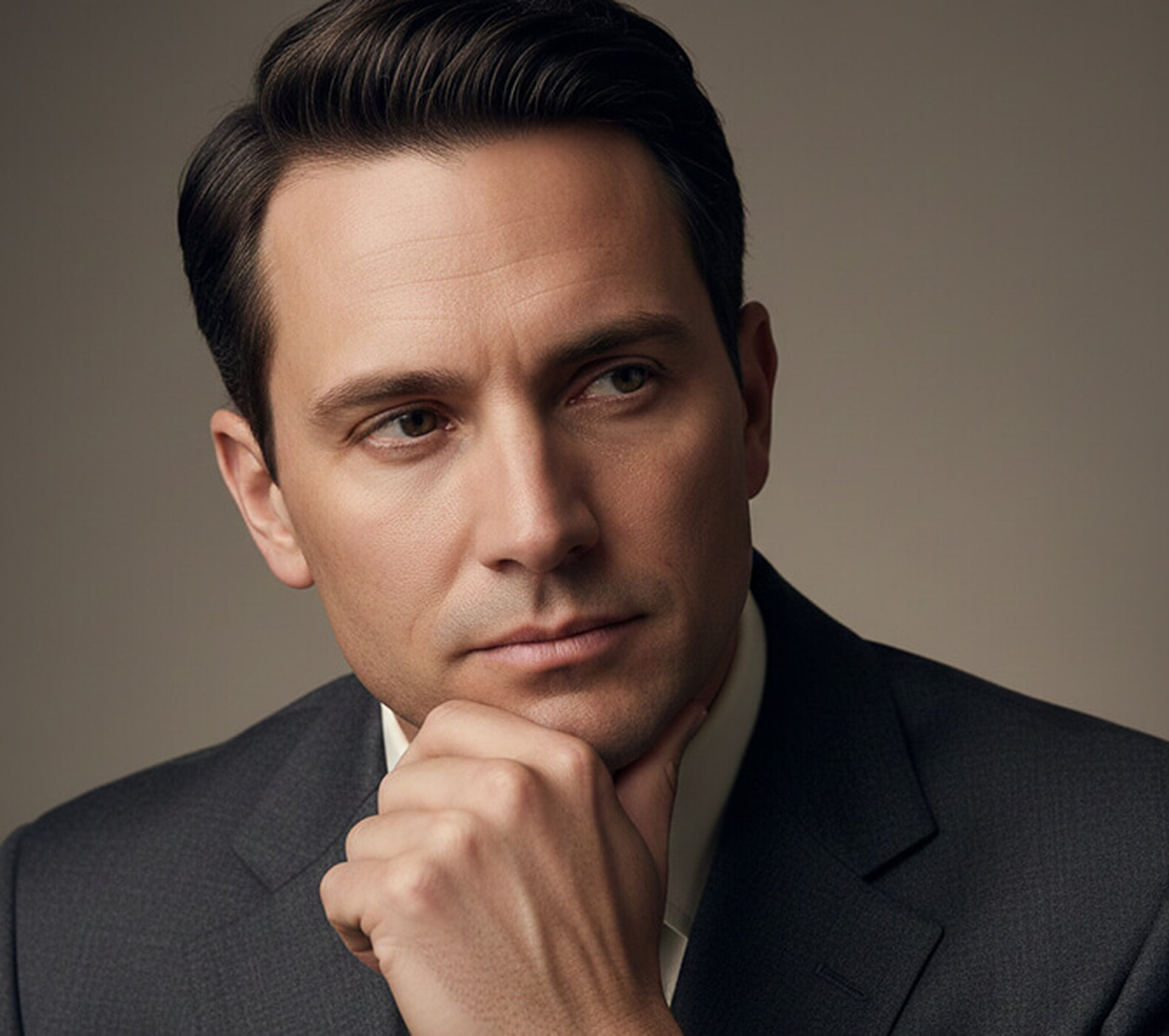 Close-crop portrait of a man in a grey suit resting a loosely curled hand against his chin and jaw, looking slightly off-camera against a warm neutral backdrop, a thoughtful corporate portrait pose.