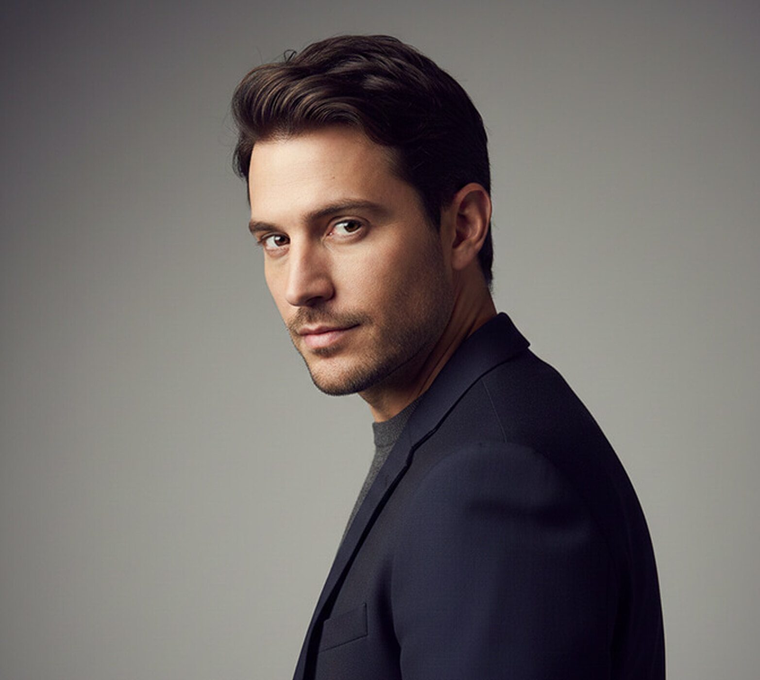 Man in a navy jacket over a grey crew-neck turned away from the camera with his head rotated back to meet the lens, shot against a grey backdrop, a dynamic corporate portrait pose.
