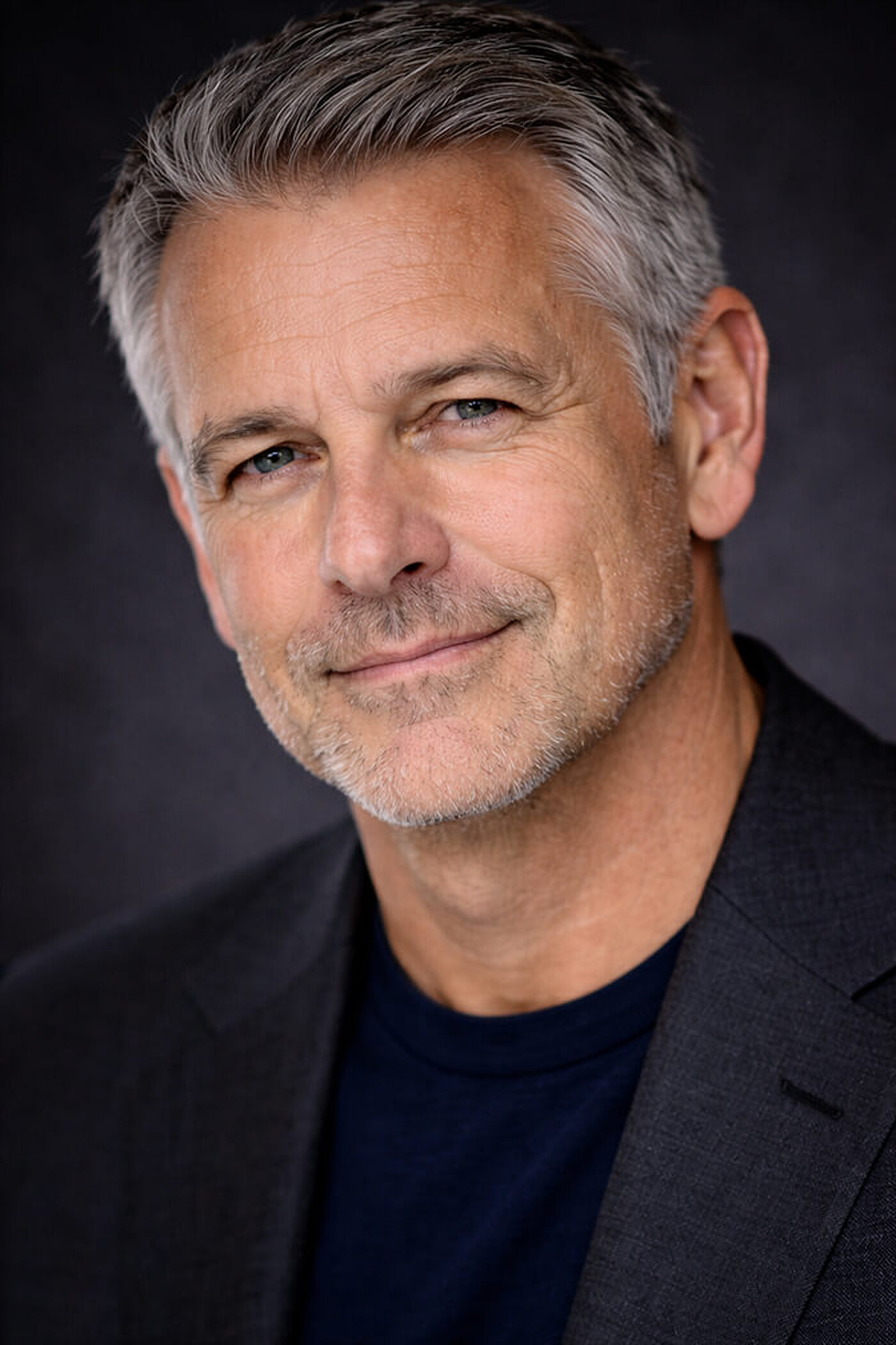 Silver-haired man in a charcoal blazer over a navy t-shirt in a tight head-and-shoulders crop, head tilted slightly with a warm closed-mouth smile against a dark backdrop, a LinkedIn profile picture pose.