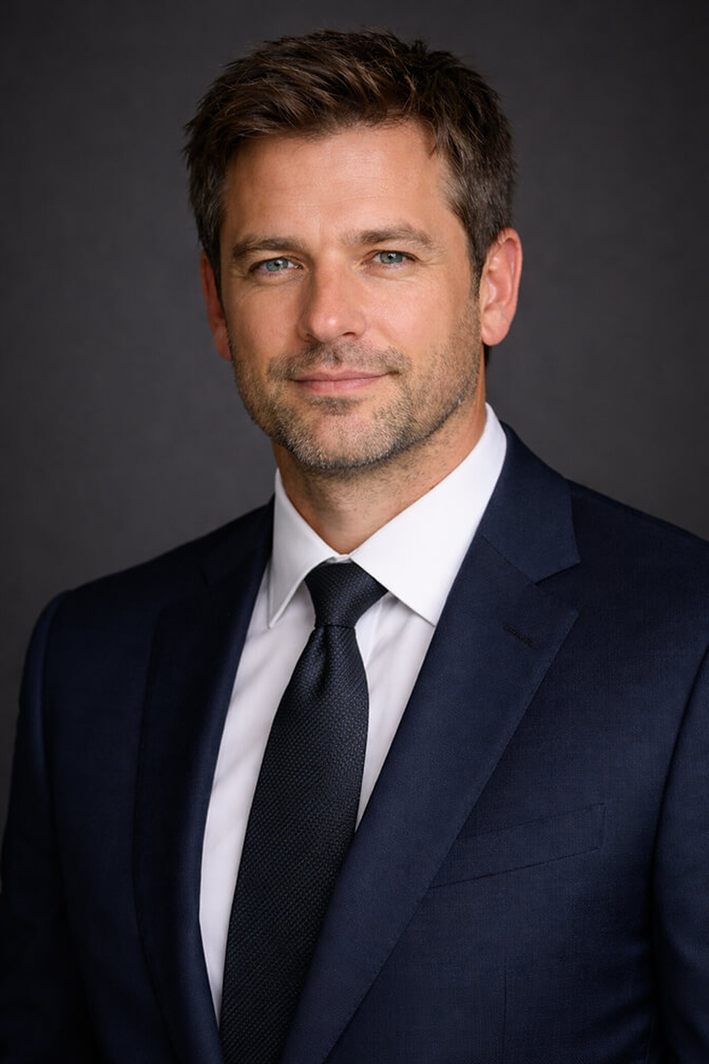 Man in a navy suit, white shirt and dark navy tie facing the camera straight on with a neutral closed-mouth expression against a dark grey backdrop, an authoritative LinkedIn profile picture pose.