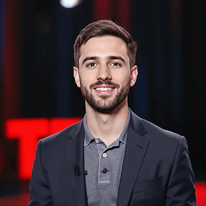 AI-generated headshot of a man with short brown hair, wearing a dark blazer and polo shirt, standing confidently in front of a TED Talk stage backdrop with vibrant lighting.