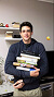 Smiling man holding a stack of books standing in an indoor setting with soft background.