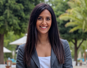 Smiling woman in a gray blazer standing outdoors with trees and a blurred background.