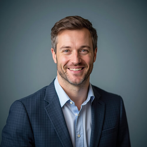 AI-generated professional headshot of a man with brown hair and short beard wearing dark blue patterned suit jacket over light blue shirt against clean gray background.