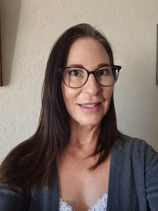 Before photo of a smiling woman wearing glasses and a grey cardigan, captured indoors with natural light.
