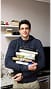 Smiling man holding a stack of books standing in an indoor setting with soft background.