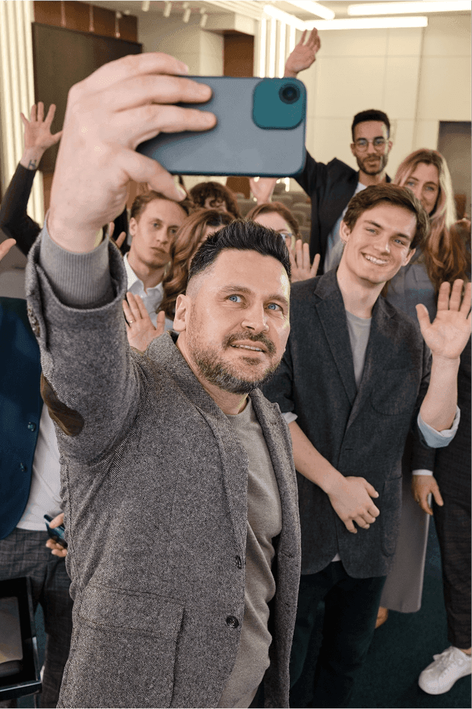 Group selfie taken at arm's length with a phone, where the person holding the camera is closest to the lens and appears noticeably larger than everyone standing behind them.