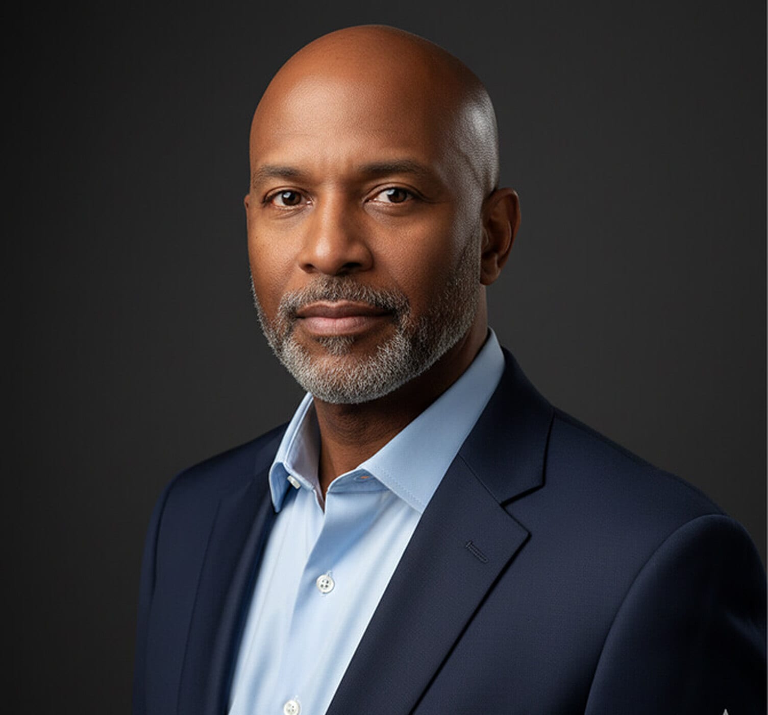 Bald man with a grey beard in a navy suit and light blue shirt with his shoulders dropped and relaxed away from his ears, photographed against a dark backdrop, showing relaxed posture in a corporate headshot.