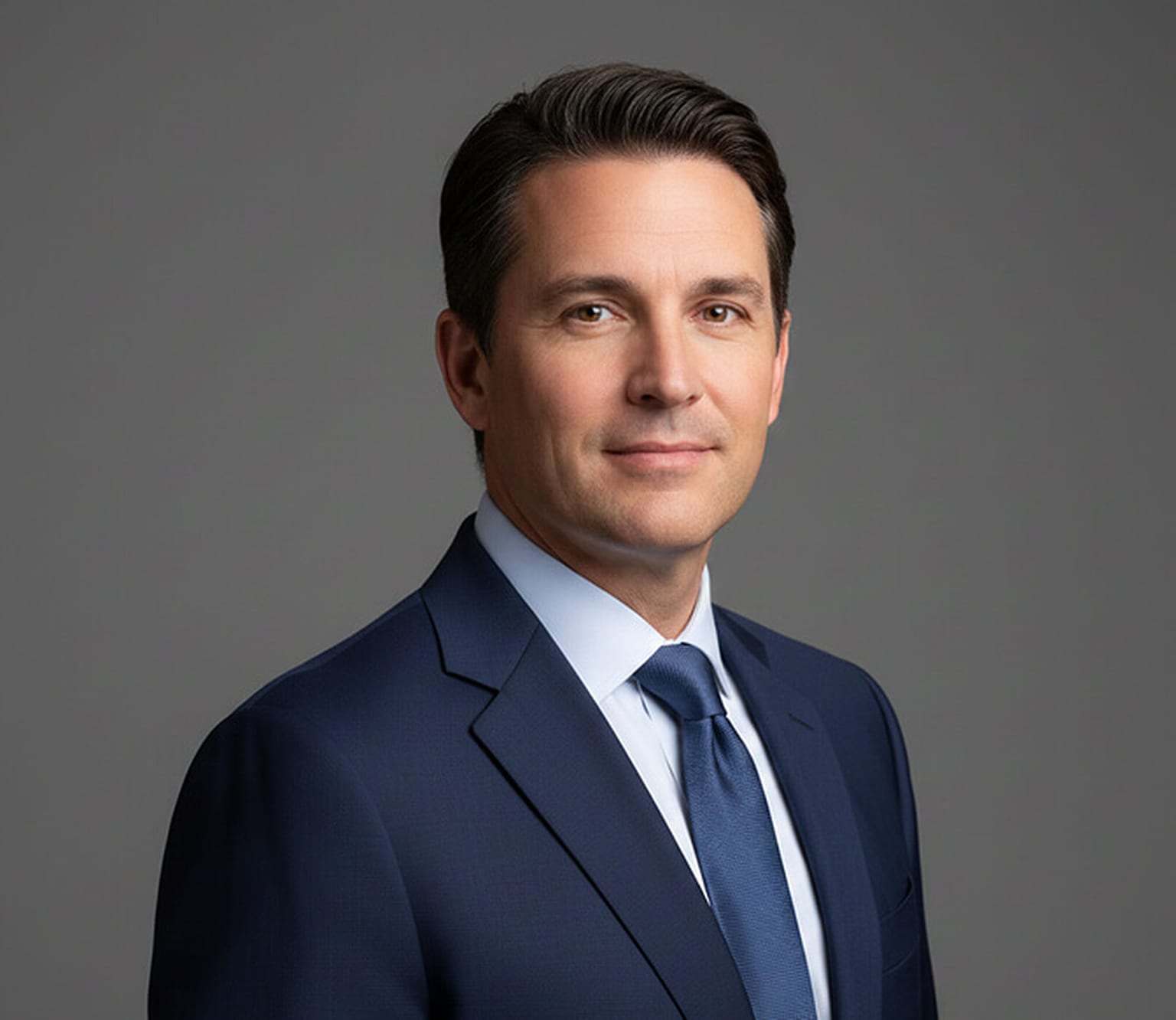 Executive in a navy suit, light blue shirt and blue tie turned at a three-quarter angle with his head level and a faint closed-mouth smile against a mid-grey backdrop, a classic senior leadership headshot pose.