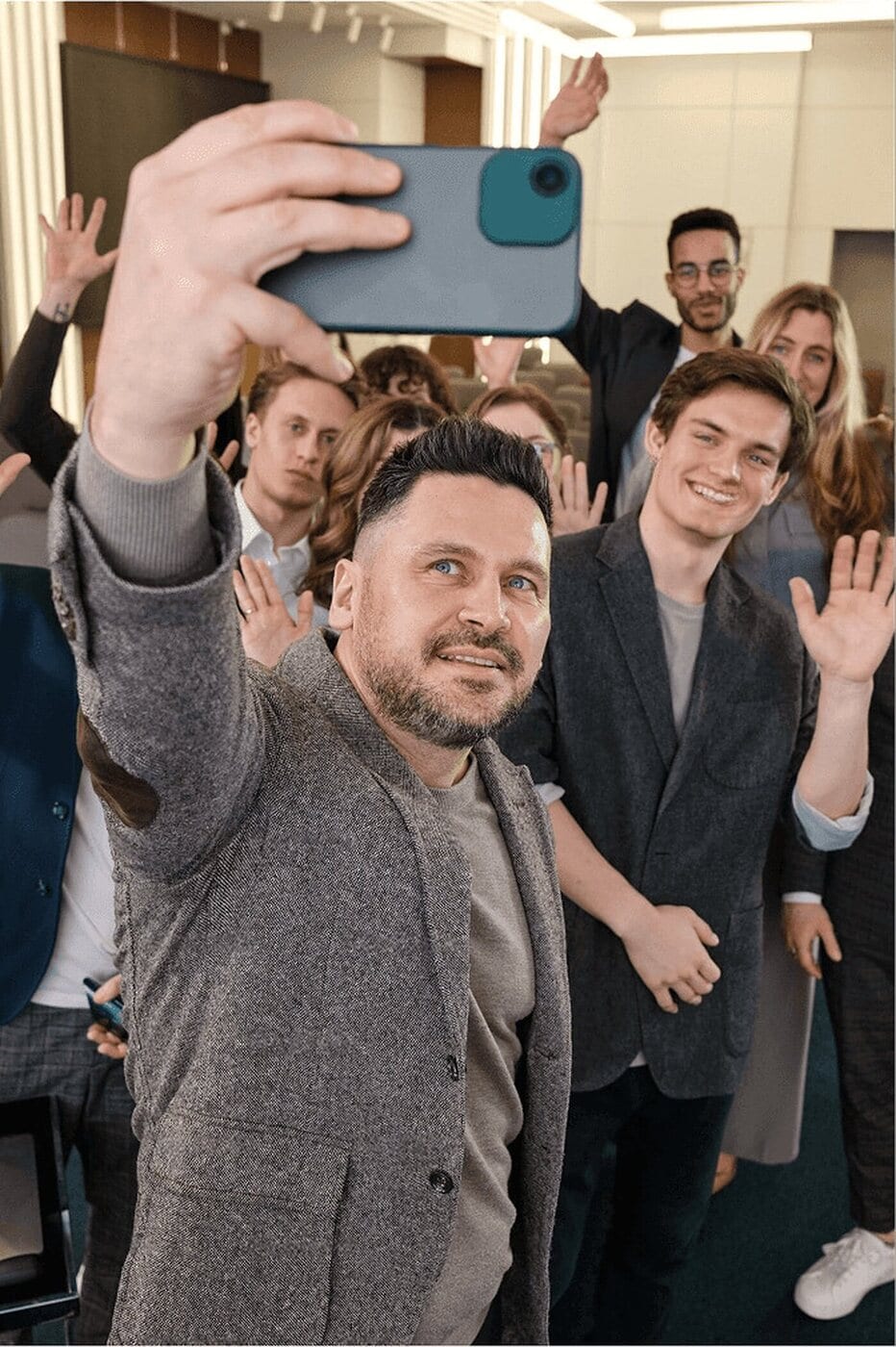 Nine colleagues crowded into an arm's-length phone selfie in an office, several waving and leaning in from the back of the group.
