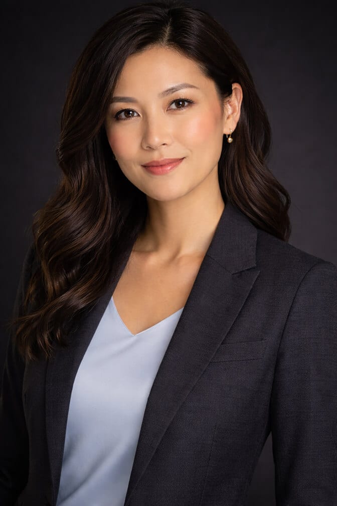 Professional AI headshot of a woman in a charcoal blazer and light blue top against a dark studio background