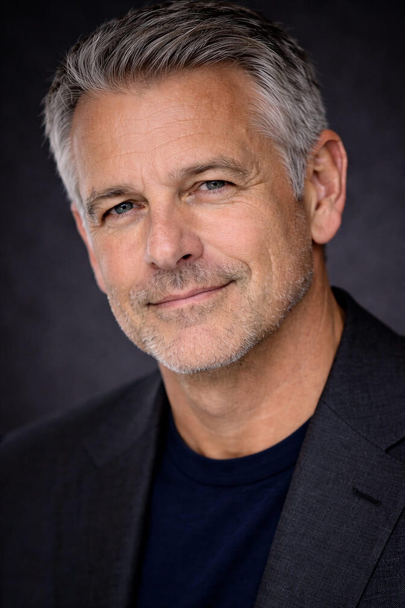Silver-haired man in a charcoal blazer over a navy t-shirt in a tight head-and-shoulders crop, head tilted slightly with a warm closed-mouth smile against a dark backdrop, a LinkedIn profile picture pose.