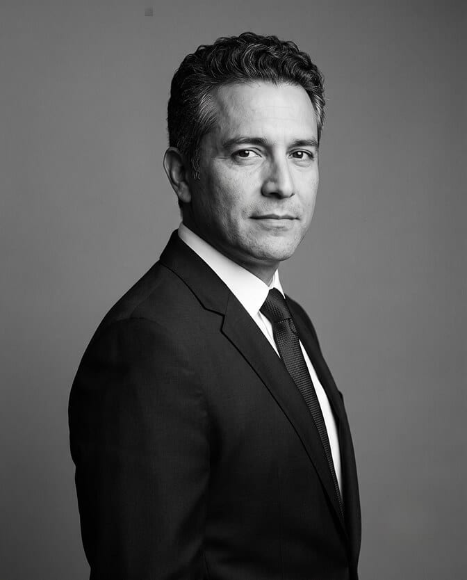 Black-and-white editorial CEO headshot: executive in a dark suit, white shirt and dark tie, body turned and face to camera against a gray gradient backdrop