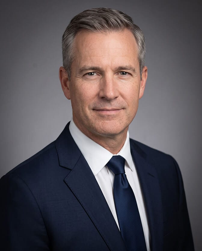 Classic boardroom male CEO headshot: gray-haired executive in a navy suit, white shirt and solid navy tie against a plain gray studio backdrop, shoulders angled and face turned to camera