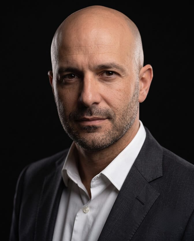 Low-key dark studio CEO headshot: bald executive with stubble in a charcoal blazer and open white shirt, lit by one strong light against a black background