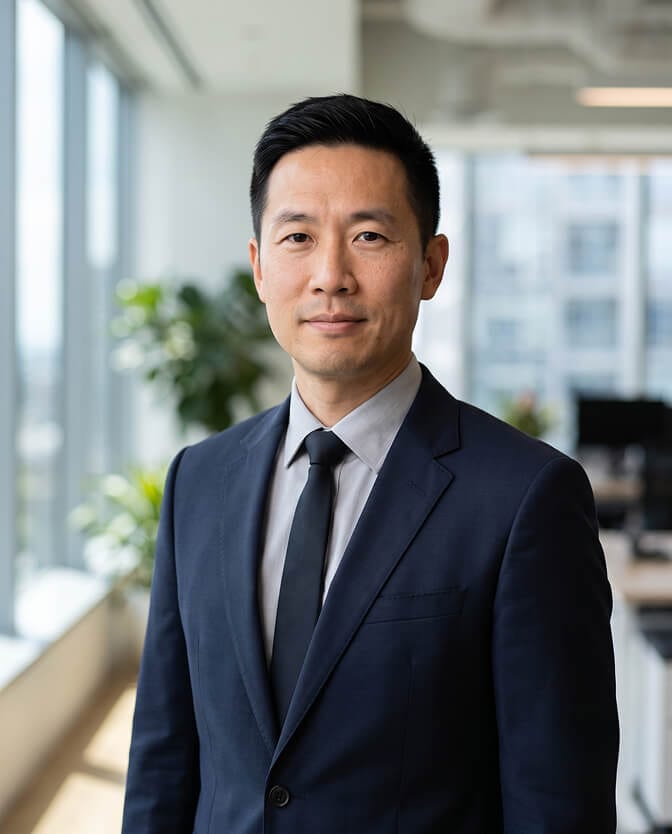 Office environment CEO headshot: executive in a navy suit and dark tie standing in a bright modern office with large windows and plants blurred behind him