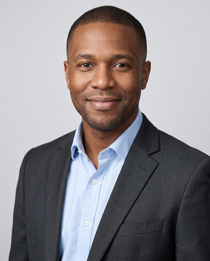 Open-collar founder CEO headshot: smiling man in a charcoal blazer over a light blue shirt with no tie, against a light gray background