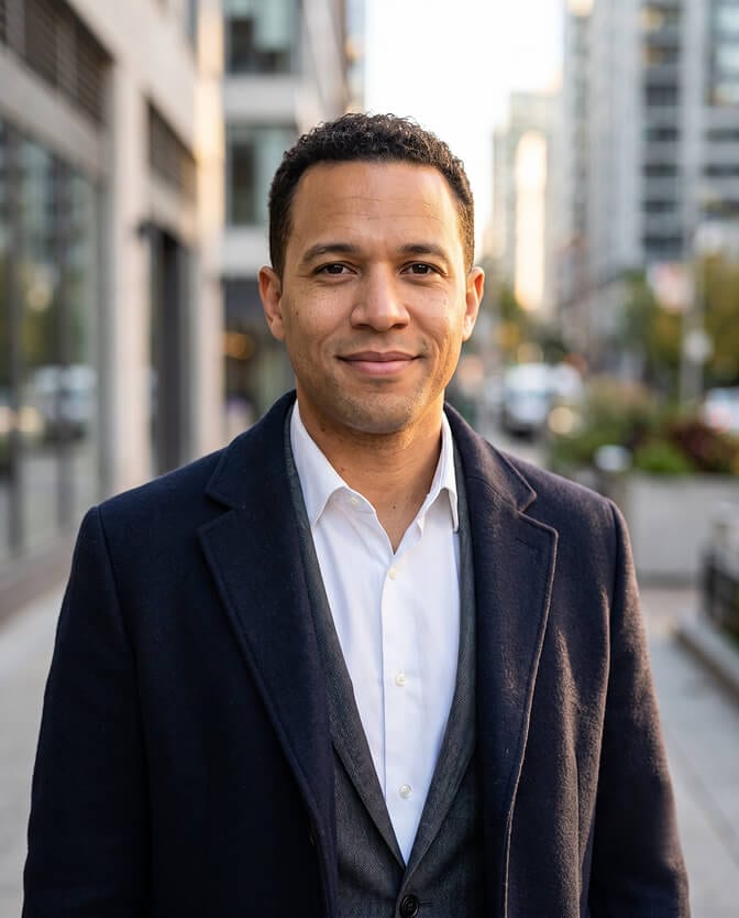 Outdoor natural light CEO headshot: smiling executive in a navy blazer and open white shirt standing on a city street with buildings and greenery softly blurred behind him