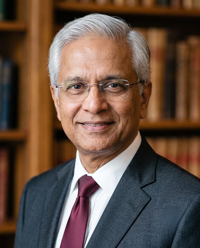 Silver-haired chairman headshot: senior executive with white hair and glasses in a charcoal suit, white shirt and burgundy tie, smiling in front of a warm wooden bookshelf