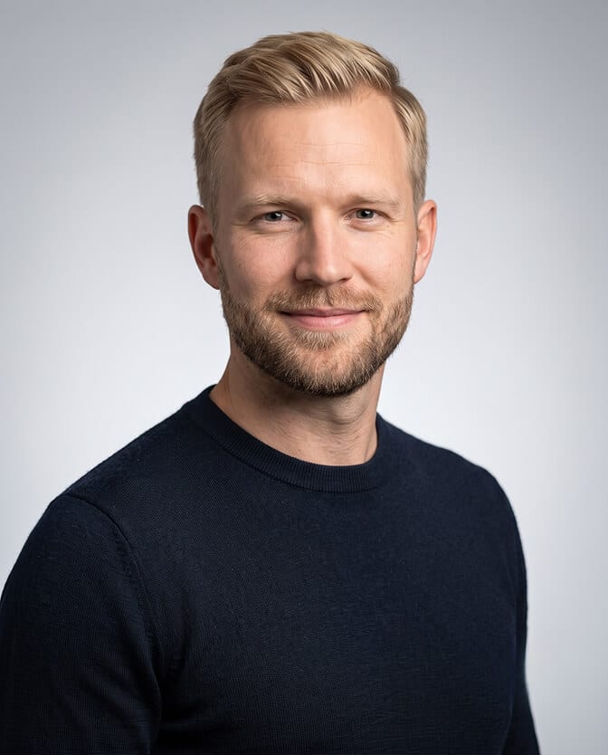 Smart-casual tech CEO headshot: smiling man with short blond hair and a trimmed beard in a fitted navy crew-neck knit against a light gray background