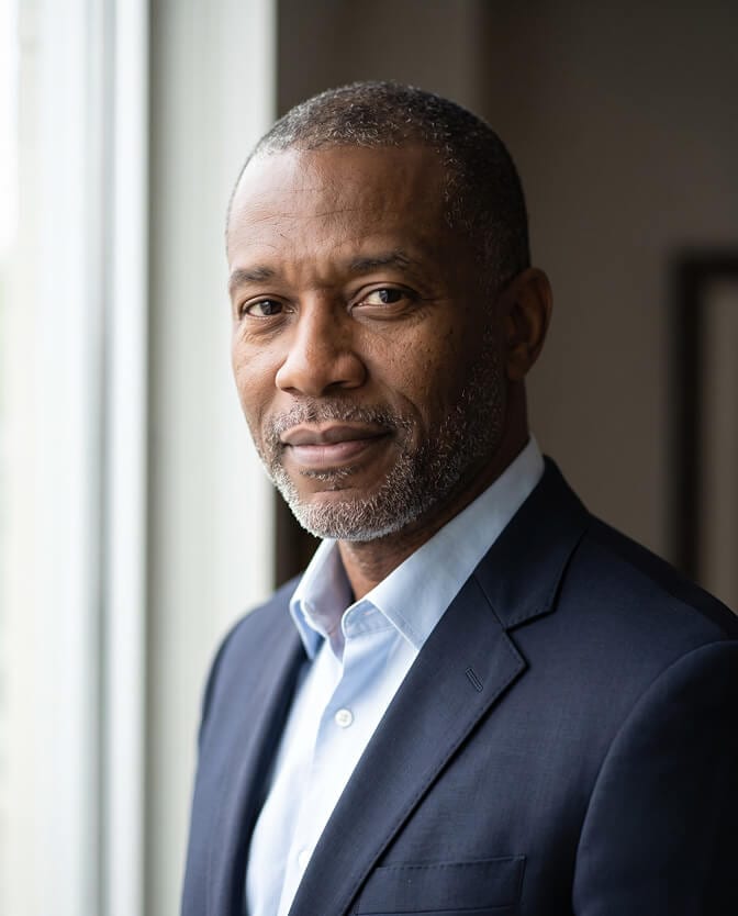 Window-light thought leader headshot: senior executive with a short gray beard in a navy suit and light blue shirt, lit softly from a window on one side with a slight turn of the body