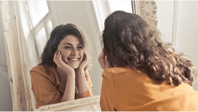 Woman looking at her reflection, illustrating the difference between the mirror image of a face that you see every day and the unflipped camera image that everyone else sees.