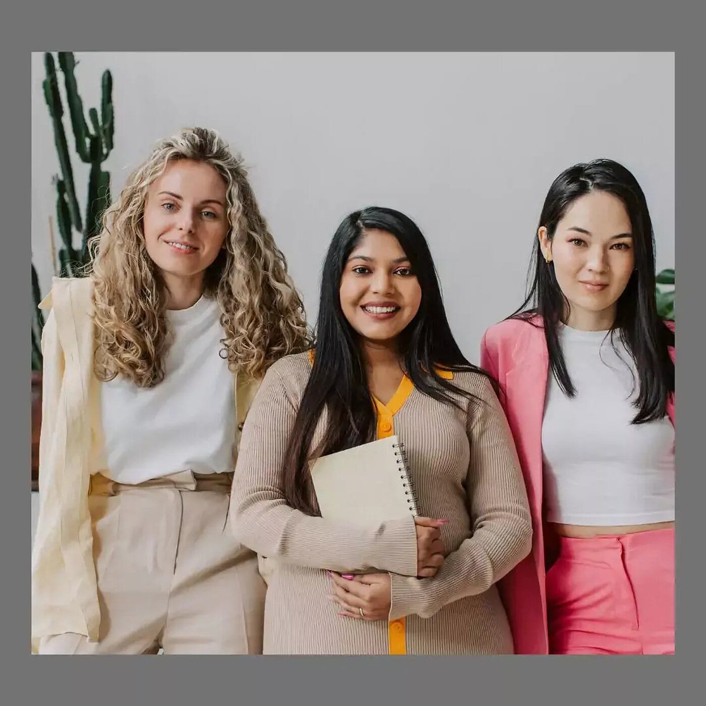 Three women standing close together in a triangular arrangement, one slightly forward holding a notebook and two behind her, in a bright neutral room.