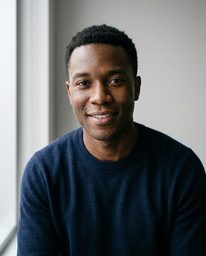 Professional iPhone style headshot of a young man in a dark navy sweater lit by a soft window from the side showing how indirect daylight creates an even flattering tone on the face