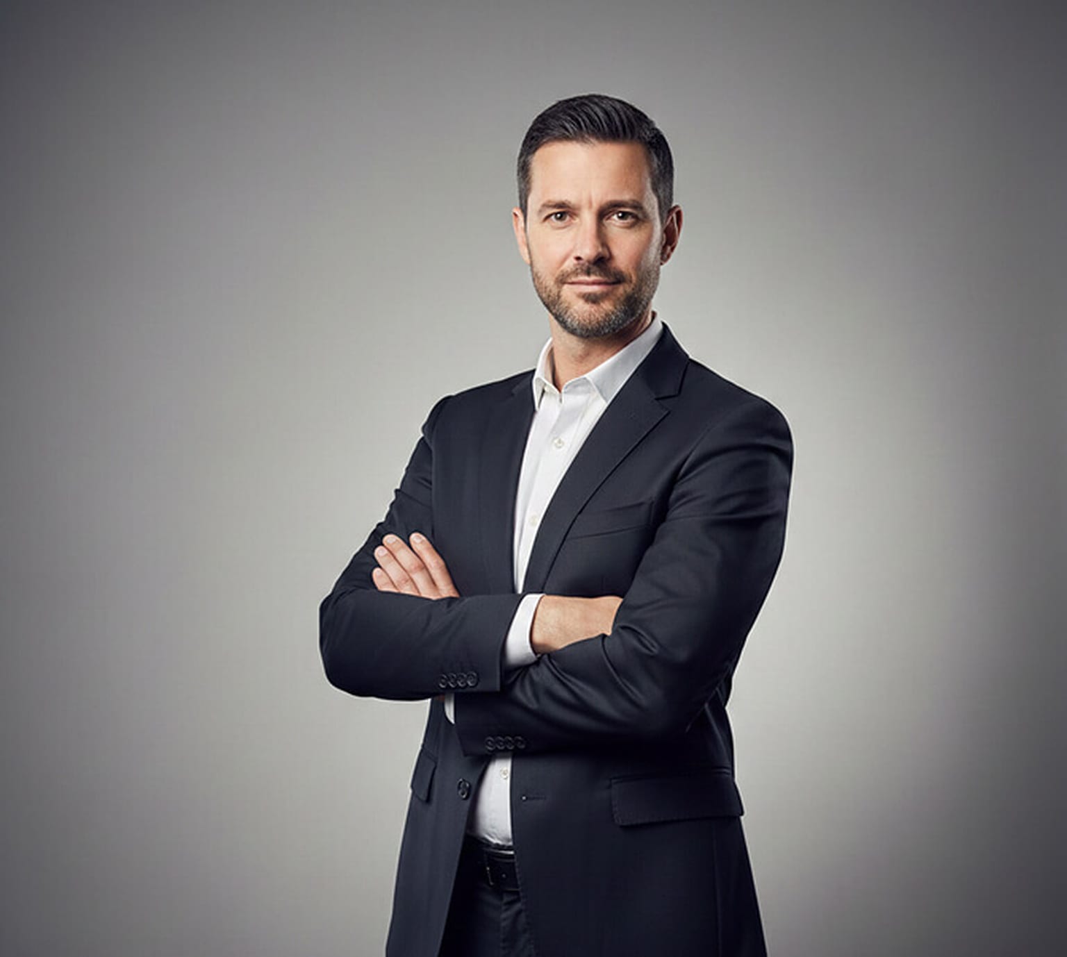 Man in a charcoal suit and white open-collar shirt standing with arms crossed at a three-quarter angle against a grey studio backdrop, a professional corporate portrait pose.