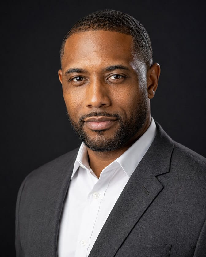 Professional AI headshot of a man in a gray blazer and white open collar shirt against a dark studio background showing a polished translation of his features through the camera
