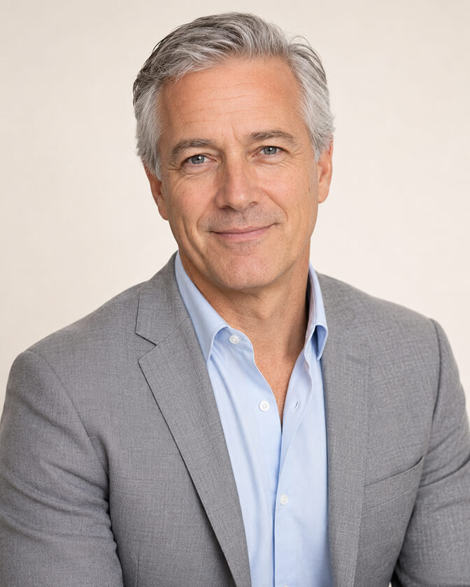 AI headshot of an executive with silver hair and a light stubble in a grey blazer over an open blue shirt, photographed against a warm neutral backdrop.