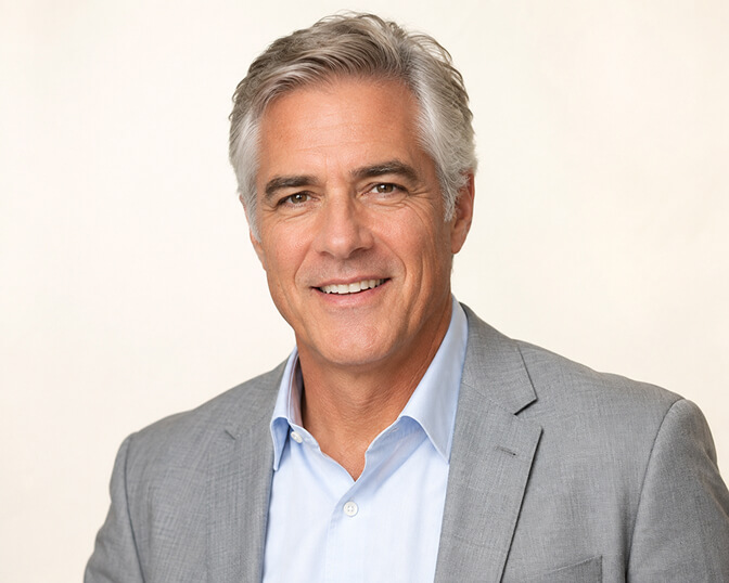 AI headshot of a smiling silver-haired executive in a light grey suit and pale blue shirt, photographed against a soft cream backdrop with bright even lighting.