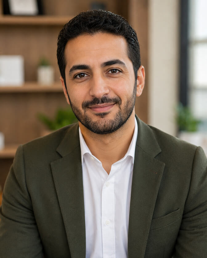 AI headshot of a bearded man in an olive green blazer over a white open-collar shirt, seated in a warm office with wooden shelving and plants blurred behind him.