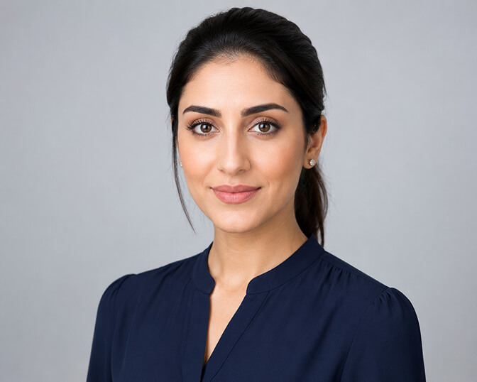 AI headshot of a woman with dark hair tied back, wearing a navy collarless blouse, photographed against a plain light grey studio backdrop with soft even lighting.