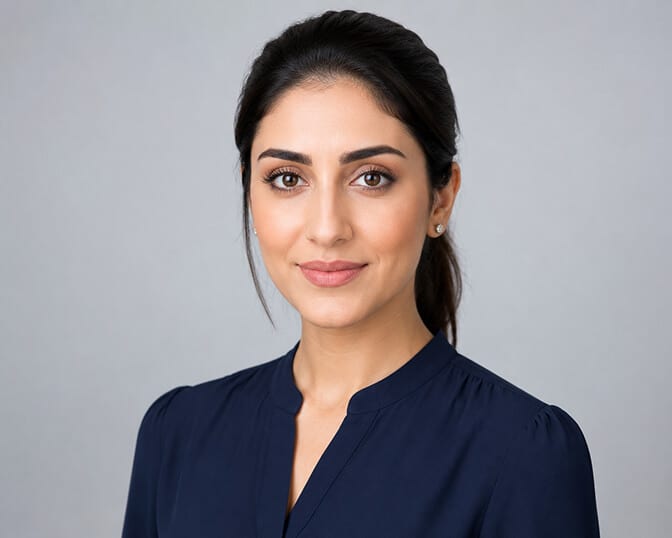 AI headshot of a woman with dark hair tied back, wearing a navy collarless blouse, photographed against a plain light grey studio backdrop with soft even lighting.