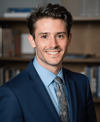 AI-generated professional headshot of a smiling young man in blue suit jacket and patterned tie, with blurred office bookshelf background