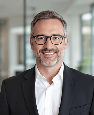 AI-generated professional headshot of a smiling middle-aged man with glasses wearing a dark suit jacket, with blurred modern office background