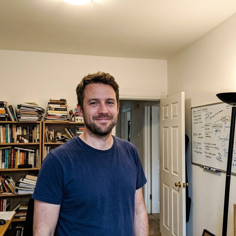 Before using the profile picture maker: a casual phone photo of a man standing off-centre in a cluttered home office, with a crowded bookshelf, an open door and a whiteboard behind him.